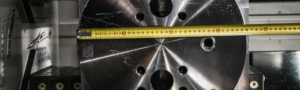 Gedrehtes Metallteil mit Bohrungen, darüber ein gelbes Bandmaß